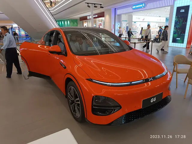 The image shows an orange car on display in a showroom, surrounded by a group of people standing on...