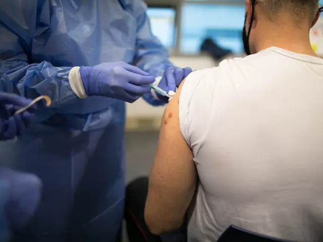 The image shows a man in a white shirt getting a vaccine from a doctor wearing a blue dress and...