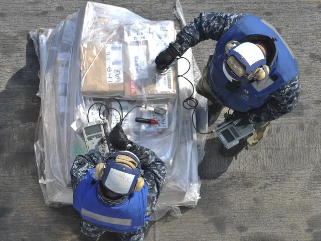 The image shows two men in blue uniforms and helmets working on a large piece of equipment, which...