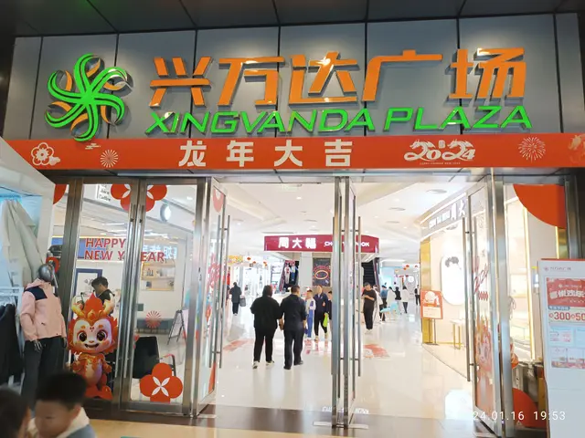The image shows the entrance to a shopping mall with people walking through it. On the left side of...