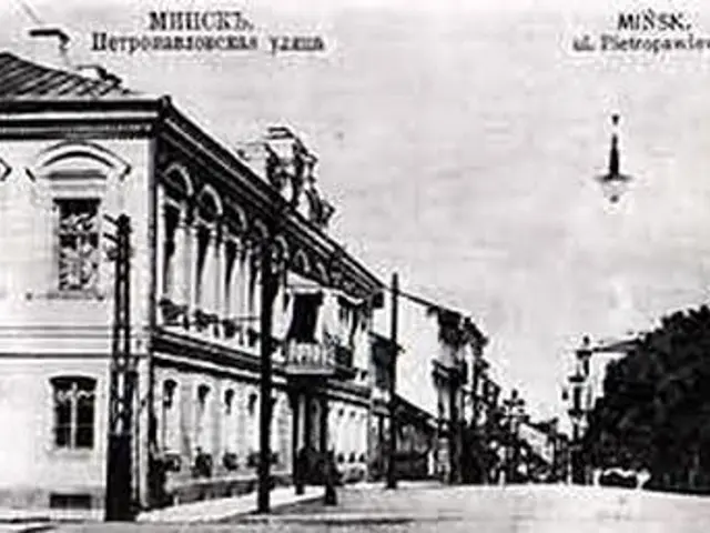 The image shows a black and white photo of a city street with buildings, street poles, street...