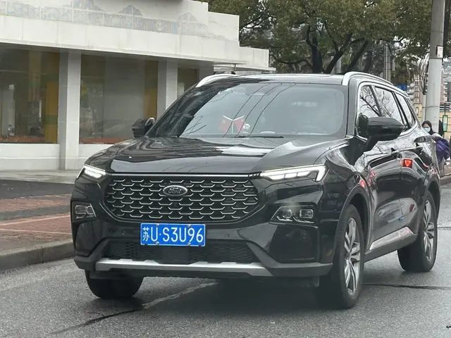The image shows a black 2020 Ford Explorer SUV driving down a city street, with a person wearing a...