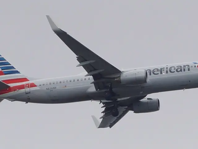 The image shows an American Airlines plane flying in the sky with its landing gear down, its wings...