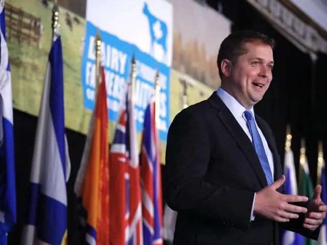 The image shows a man in a suit and tie standing at a podium with a microphone in front of him,...