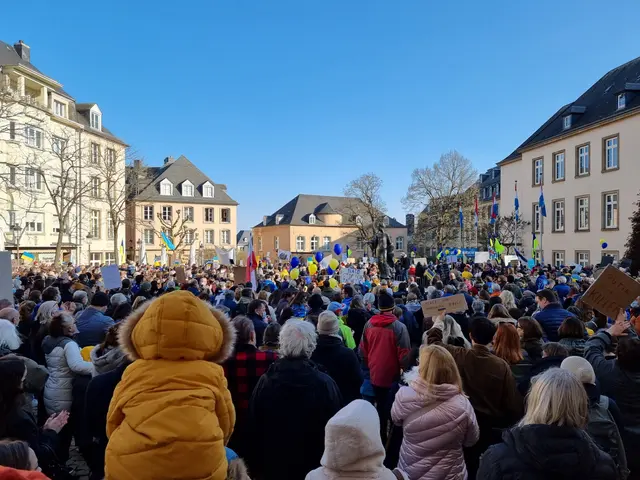 The image shows a large crowd of people standing in front of a building, holding placards and...