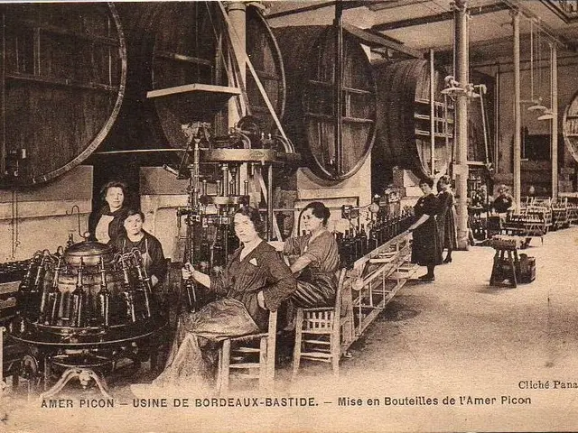 The image shows an old black and white photo of a group of women working in a factory. There are...
